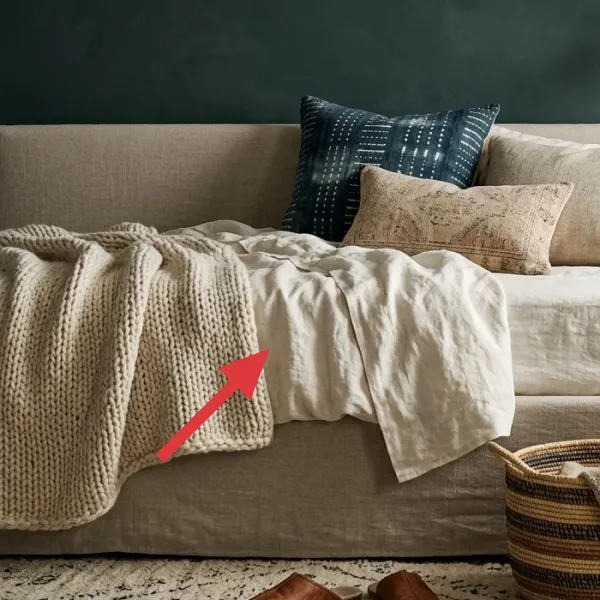 Close-up of layered bedding: off-white linen duvet, chunky knit throw, and two decorative pillows (blue patterned and beige textured).