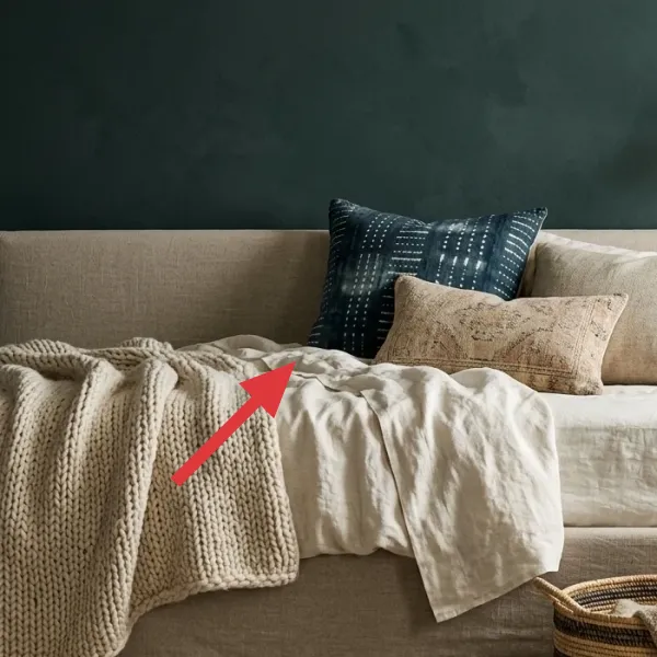 Close-up of a bed with layered linen duvet, chunky knit throw, and an assortment of decorative pillows in neutral and blue tones.