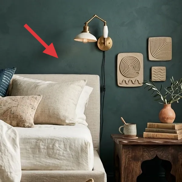 A brass-finished plug-in wall sconce with a white shade and pivoting arm, mounted on a dark teal wall above a bed.